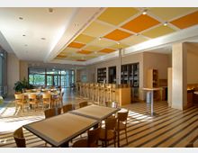 Großer, moderner Café- oder Restaurantbereich mit Holzmöbeln, gestreiftem Holzboden und einer langen Bar mit Barhockern. Die Decke ist mit orange- und gelbfarbenen Akzentflächen gestaltet, seitlich befinden sich große Fenster, die Tageslicht hereinlassen.