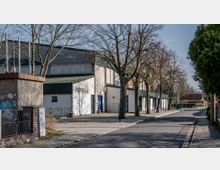 Straßenansicht einer Stadthalle mit weißer Fassade und grünem Dach, flankiert von kahlen Bäumen. Im Vordergrund ein Gehweg mit Laternen, im Hintergrund eine ruhige Wohnstraße mit einzelnen Häusern.