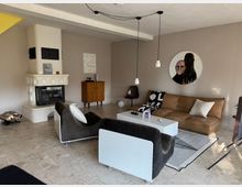 Modern eingerichtetes Wohnzimmer mit cremefarbenem Kamin, braunem Ledersofa, zwei Sesseln, hellem Couchtisch und Kunstwerken an den Wänden. Der Raum hat einen hellen Fliesenboden und ist minimalistisch dekoriert.