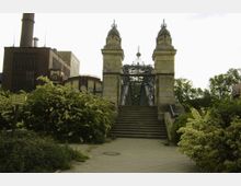 Ein historisches Brückenportal mit steinernen Säulen und dekorativen Spitzen führt über eine Treppe zu einer schmiedeeisernen Brücke. Im Hintergrund sind Industriebauten aus Backstein und moderner Architektur sowie umgebende Grünflächen zu sehen.