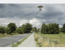 Eine ländliche Straße mit einem Gehweg und einzelnen Straßenlaternen führt durch eine grüne Landschaft. Auf der rechten Seite befinden sich Wiesen mit vereinzelten Zäunen, links stehen dichte Bäume, und der Himmel ist von dunklen Wolken bedeckt.