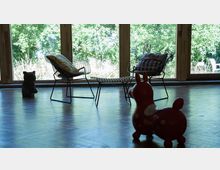 Innenraum mit Holzboden, großer Fensterfront mit Blick ins Grüne. Im Vordergrund ein rotes Spielzeug in Tierform, zwei Drahtstühle mit Kissen sowie eine kleine Tierfigur aus Stein.