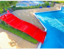 Ein Freibad mit einer roten Wasserrutsche, die in ein großes, blaues Becken führt. Im Hintergrund sind gepflasterte Gehwege, kleinere runde Wasserbecken, Sitzbänke und grüne Büsche zu sehen.