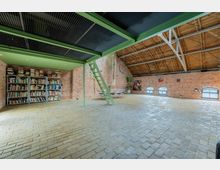 Großzügiger, offener Raum in einem Loft-Stil mit Backsteinwänden, Holzdachkonstruktion und großen, halbkreisförmigen Fenstern. Im Vordergrund befinden sich Bücherregale, eine grüne Metalltreppe führt zu einer Zwischendecke, im hinteren Bereich stehen Tische und Stühle sowie Pflanzen und Wanddekorationen.