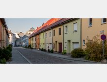 Straßenansicht mit Reihenhäusern in unterschiedlichen Pastellfarben, flankiert von einem gepflasterten Gehweg und Bäumen. Im Hintergrund sind moderne Gebäude zu sehen, ein Verkehrsschild weist ein Halteverbot aus.