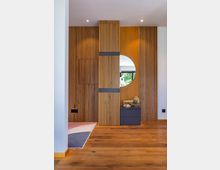 Innenraumansicht mit einer Holzvertäfelung aus vertikalen Streifen, einem runden Spiegel und einer schwarzen Kommode mit Dekorationen wie Blumen und Bilderrahmen. Der Boden besteht aus warmem Holzparkett, und ein Teil eines Teppichs ist am linken Rand sichtbar.