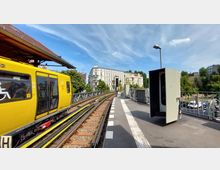 Eine oberirdische U-Bahn-Station mit einem gelben Zug auf Gleisen, die sich durch eine städtische Umgebung mit historischen weißen Gebäuden und grünen Bäumen schlängeln. Der Bahnsteig ist mit einem Blindenleitsystem ausgestattet, und im Hintergrund ist ein klarer blauer Himmel zu sehen.