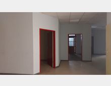 Ein leerer Innenraum mit hellgrauen Wänden und rotem Türrahmen. Es sind mehrere offene Türen zu sehen, eine führt zu einem Bereich mit Glasfenstern und Holztüren im Hintergrund. Die Decke ist mit weißen Rasterplatten und einer Leuchtstofflampe ausgestattet.