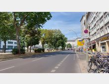 Städtische Straße mit breiten Gehwegen, auf denen Fahrräder geparkt sind, sowie einer mehrspurigen Fahrbahn mit Bäumen auf der linken Seite. Rechts befinden sich Mehrfamilienhäuser und Geschäfte, darunter ein Postshop, und mehrere Verkehrsschilder.