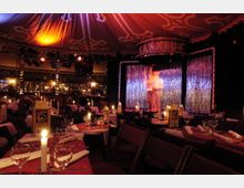 Ein festlich dekorierter Varieté-Saal mit gedeckten Tischen, Kerzenlicht und einer kleinen Bühne, die mit glitzernden Vorhängen und bunten Scheinwerfern beleuchtet ist. Im Hintergrund ist der Schriftzug „Kat Klub“ über der Bühne zu erkennen.