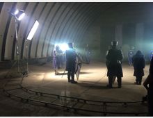 Personen in mittelalterlicher Kleidung mit Rüstungen und Schilden stehen in einer Halle mit gebogenen Wänden und industriellem Charakter. Im Vordergrund sind Schienen und Studiobeleuchtung zu sehen, was auf eine Film- oder Fotoszene hindeutet.