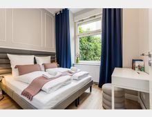 Helles Schlafzimmer mit Doppelbett, gepolstertem Kopfteil und weißer Bettwäsche, dazu ein kleiner weißer Schreibtisch und Hocker. Großes Fenster mit dunkelblauen Vorhängen und Blick ins Grüne, darunter ein Heizkörper.