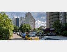 Blick auf eine Wohnsiedlung mit mehreren Hochhäusern, umgeben von Bäumen und Grünflächen. Im Vordergrund sind geparkte Autos und ein Gehweg zu sehen; der Himmel ist teils bewölkt.