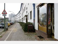 Straßenansicht mit einer langen weißen Wohnhausfassade im rechten Bildbereich, einem Friseursalon mit Schaufenster im Vordergrund und parkenden Autos entlang des Gehwegs auf der linken Seite. Im Vordergrund steht ein Verkehrsschild mit der Aufschrift „Straßenschäden“.