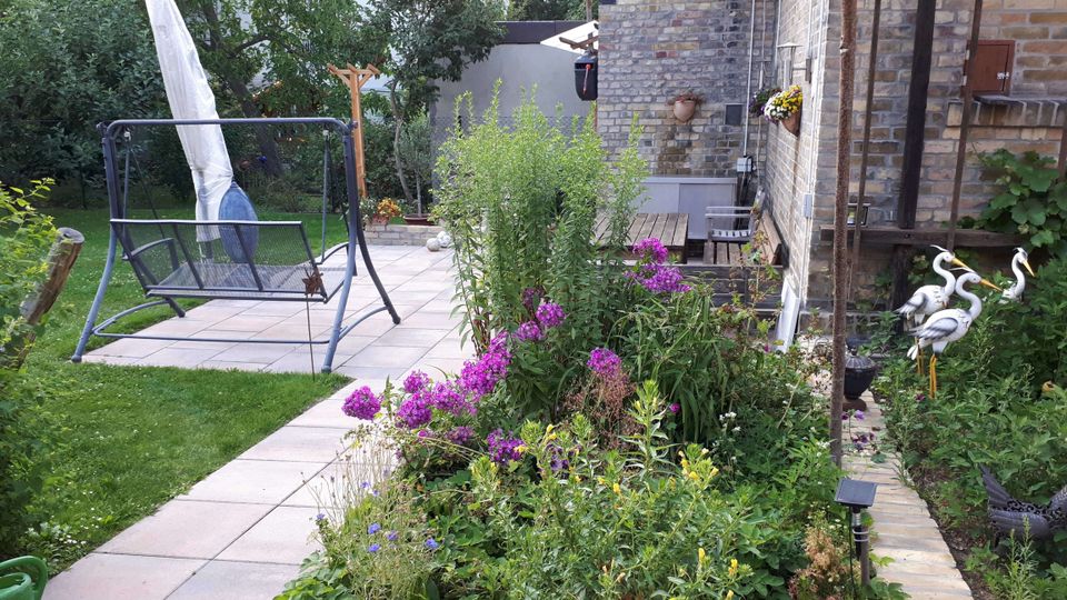 Gepflegter Hausgarten mit gepflastertem Weg und kleiner Terrasse mit Gartenschaukel und geschlossenem Sonnenschirm. Rechts steht ein Backsteinhaus mit Rankpflanzen, daneben ein dichtes Blumenbeet mit lila Blüten und Gartenfiguren am Rand.