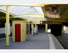 Ein U-Bahnsteig mit gelben Säulen und einem überdachten Glasdach. Zu sehen sind ein gelbes Backsteingebäude mit roten Türen, eine Uhr, ein Fahrplananzeiger mit Abfahrtszeiten sowie Gleise auf der rechten Seite.