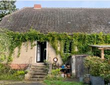 Ein älteres Einfamilienhaus mit Satteldach, dessen Fassade teils von grünem Efeu überwuchert ist. Vor dem Eingangsbereich führen einige Stufen zur offenen Haustür, daneben stehen Blumentöpfe und kleinere Gartenobjekte, während rechts ein Holzschuppen sichtbar ist.