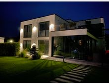 Modernes zweistöckiges Wohnhaus bei Nacht, mit weißer Fassade, großen Glasfronten und einem bepflanzten Balkon. Im Vordergrund ist ein gepflegter Garten mit Rasen, Büschen und einem gepflasterten Weg zu sehen, beleuchtet durch Außenlampen.