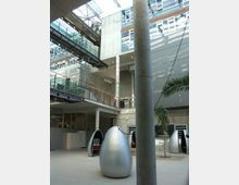 Moderne Innenansicht eines Bürogebäudes mit Betonwänden, großen Glasflächen und einem offenen Deckensystem. Im Vordergrund stehen mehrere silberne, eiförmige Sitzkapseln, während im Hintergrund Brücken und Geländer verschiedene Ebenen verbinden.