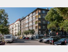 Straßenansicht eines Wohngebäudes mit fünf Stockwerken, dessen Fassade in dunklen Grautönen gestaltet ist, daneben ein angrenzendes helles Gebäude. Vor den Gebäuden stehen parkende Autos, Wohnwagen und Bäume, die teilweise Schatten spenden. Straßenschilder und Laternen sind sichtbar.