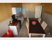 Rustikale Küche mit gelben Wänden, weiß-blauen Hängeschränken, Spüle und kleiner Arbeitsfläche. Im Raum steht ein dunkler Esstisch mit vier Stühlen, auf dem ein roter Teller liegt.