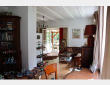 Wohnzimmer mit Holzboden und verschiedenen Möbeln, darunter ein Schreibtisch, eine braune Ledercouch, ein Bücherregal und ein Beistelltisch. Im Hintergrund öffnet sich eine Tür zu einer überdachten Terrasse mit Blick ins Grüne.