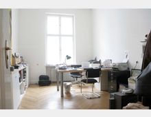 Helles Büro mit Holzboden, großen weißen Wänden und einem großen Fenster in der Mitte. Der Raum enthält einen Schreibtisch mit Büromaterialien, Stühlen, Regalen an der linken Wand und einem Drucker auf der rechten Seite.