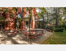 Außenterrasse mit rot-braunen Tischen und Stühlen vor einem Backsteingebäude mit großen Glasfenstern. Im Hintergrund sind ein gläserner Anbau und umgebende Bäume sichtbar.