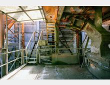 Industrielle Anlage mit rostigen Metalltreppen, Geländern und Rohrleitungen, umgeben von verwitterten Wänden und einer teils durchlässigen Überdachung. Die Szenerie zeigt Gebrauchsspuren wie Rost und abgeblätterte Farbe.