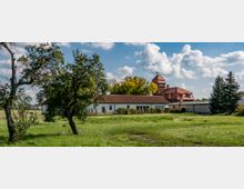 Weitläufige Wiese mit vereinzelten Bäumen im Vordergrund, dahinter ein Gebäudekomplex bestehend aus einem weißen länglichen Haus mit rotem Ziegeldach und einem angrenzenden Gebäude mit einem Turm und Holzverkleidung. Klarer Himmel mit lockeren weißen Wolken.