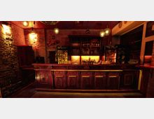 Gemütliche Bar mit einer langen Holztheke und detaillierten Verzierungen, dahinter ein Regal mit Flaschen und Gläsern, beleuchtet durch warmes, gelbliches Licht. Die Wände bestehen aus rohem Mauerwerk, und an der Decke hängen runde Lampen.
