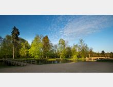 Parklandschaft mit einem breiten Kiesweg im Vordergrund, einer hölzernen Brücke, die über einen kleinen Fluss führt, und grünen Bäumen im Hintergrund unter einem klaren, blauen Himmel mit weißen Wolken.