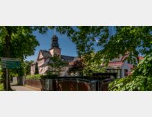 Historisches Gebäude mit rosafarbener Fassade und Uhrturm, umgeben von Bäumen und Hecken. Im Vordergrund ein Gehweg mit Wegweisern und ein metallisches Zugangstor.