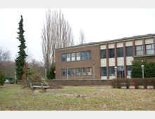 Gebäude aus rotem Backstein mit rechteckigen Fenstern und beigefarbenen Akzenten, umgeben von einer Grünfläche mit kahlen Bäumen und Büschen. Im Vordergrund steht eine Parkbank, und der Boden ist mit herabgefallenem Laub bedeckt.