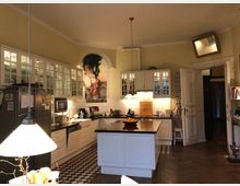 Helle, klassische Küche mit cremefarbenen Wänden und hohen Decken, einer zentralen Kochinsel mit dunkler Arbeitsplatte und weißen Einbauschränken mit Glasfronten. Der Boden besteht aus gemusterten Fliesen und Fischgrät-Holzparkett, im Hintergrund hängt ein großes Gemälde unter einem Rundbogen.