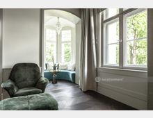 Wohnraum mit modernem Design, bestehend aus einem grünen Polstersessel, einem Hocker und einem bogenförmigen Erker mit großen Fenstern. Im Erker befindet sich eine blaue gepolsterte Sitzbank mit Kissen, ein kleiner goldfarbener Beistelltisch und eine Hängelampe.