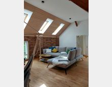 Wohnzimmer in einem Dachgeschoss mit Holzdecke und mehreren Dachfenstern, einem Holzfußboden, einer grauen Eckcouch mit bunten Kissen, einem Beistelltisch und einer Ziegelwand im Hintergrund. Ein Hängesessel sowie ein Heizkörper sind ebenfalls sichtbar.