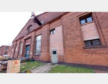 Ziegelbau mit großen, hohen Fenstern und einem schmalen Eingangstor, vermutlich Teil eines Industrie- oder Lagergebäudes. Im Vordergrund befinden sich gestapelte Baumaterialien und eine kleine Grünfläche.