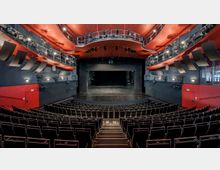 Innenansicht eines modernen Theatersaals mit ansteigenden Sitzreihen, einer schwarzen Bühne und einer roten Decke. Der Saal ist mit Beleuchtungstechnik ausgestattet, und die Wände sind in Schwarz und Rot gestaltet.