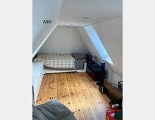 Dachgeschosszimmer mit schrägen weißen Wänden, einem Einzelbett mit karierter Bettdecke, hellem Holzboden und einem Dachfenster auf der rechten Seite. Möbel wie eine niedrige Kommode, ein Nachttisch und ein Flachbildfernseher stehen im Raum.