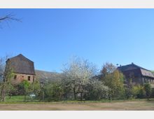 Ländliche Szenerie mit alten Gebäuden aus Ziegelsteinen und Schindeldächern, umgeben von blühenden Bäumen und Büschen. Im Vordergrund ist ein unbefestigter Boden zu sehen, während im Hintergrund ein Zaun und eine grüne Vegetation die Szene einrahmen.