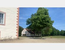 Ein sandiger Platz mit einem Baum in der Mitte und einer Wiese im Hintergrund. Links im Bild ist ein Gebäude mit heller Fassade und rot-weißen Kanten zu sehen, weiter hinten ein weiteres, helles Gebäude mit rotem Dach.