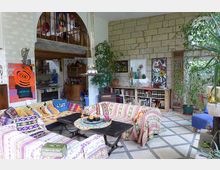 Wohnzimmer mit bunten Stoffsofas und orientalischen Mustern, dekoriert mit Kunstwerken, Pflanzen und antiken Möbeln. Im Hintergrund befindet sich eine offene Küche und eine Galerie mit Rundbogen.