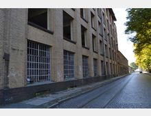 Ziegelgebäude mit mehreren Etagen und großen, vergitterten Fenstern, das entlang einer Kopfsteinpflasterstraße mit parallel verlaufenden Straßenbahnschienen steht; rechts der Straße wachsen Bäume, die Schatten spenden.