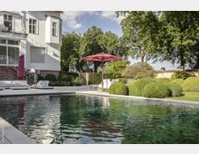 Gartenansicht mit einem Pool im Vordergrund, umgeben von gepflegten Hecken und Bäumen. An der Seite steht ein weißes Gebäude mit Backsteinsockel, außerdem sind Gartenmöbel und ein roter Sonnenschirm auf einer Terrasse zu sehen.