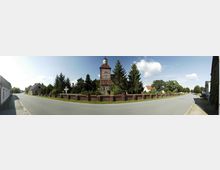 Straßenansicht mit einer historischen Fachwerkkirche in der Mitte, umgeben von einer Backsteinmauer und hohen Bäumen. Rechts und links der Straße befinden sich Wohnhäuser und kleinere Gebäude, die in eine ländliche Umgebung eingebettet sind.