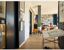 Wohnraum mit hellem Holzboden, links ein schmaler Flur mit Bildern an der Wand, im Hintergrund ein offener Bereich mit Bücherregal von Wand zu Wand, einem großen Sessel, Tisch und Dekorationen. Das Tageslicht kommt durch große Fenster mit blauen Vorhängen.