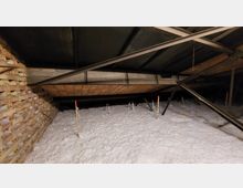 Dachbodenansicht mit offener Konstruktion aus Metallträgern, einer Ziegelsteinwand und einer dicken Schicht aus weißem Isolationsmaterial auf dem Boden. Die Decke besteht aus Dachpanelen, unterstützt durch schräge Streben.