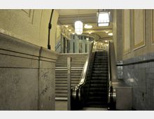 Treppenhaus in einem U-Bahnhof mit einer modernen Rolltreppe und einer danebenliegenden Steintreppe. Die Umgebung ist mit gelben und blauen Fliesen verkleidet, mit hohen Decken und geometrischen Deckenlampen.
