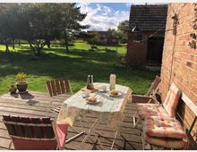 Holzterrasse mit einem Tisch, der mit einer gemusterten Tischdecke, Frühstücksgeschirr, Gebäck und einer Kaffeekanne gedeckt ist. Rund um den Tisch stehen Stühle mit gepolsterten Kissen, im Hintergrund ein Garten mit Bäumen und eine Backsteinmauer mit kleiner angelehnter Gartenlaube.